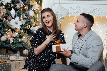 Cute romantic lovers. Happy pair hold a New Year's gift. New year eve, ornated fir tree. Winter holiday and love concept. Happy couple by the fireplace near the Christmas tree.