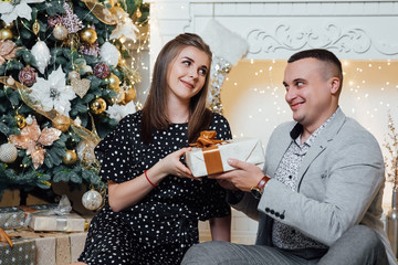 Cute romantic lovers. Happy pair hold a New Year's gift. New year eve, ornated fir tree. Winter holiday and love concept. Happy couple by the fireplace near the Christmas tree.