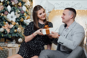 Young couple staying at home together on holiday evening sitting in front of christmas tree and hugging. Cute romantic lovers. Happy pair hold a New Year's gift.