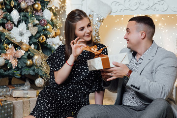 Young couple staying at home together on holiday evening sitting in front of christmas tree and hugging. Cute romantic lovers. Happy pair hold a New Year's gift.