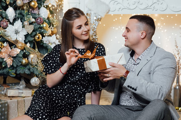 Young couple staying at home together on holiday evening sitting in front of christmas tree and hugging. Cute romantic lovers. Happy pair hold a New Year's gift.