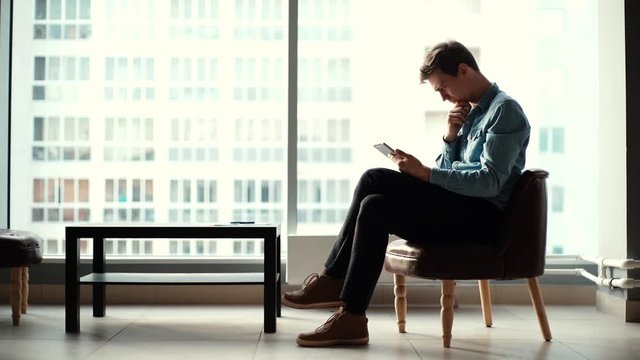 Pensive young business man sitting on chair and uses modern gadget in the office against a large window. Shooting in slow motion.