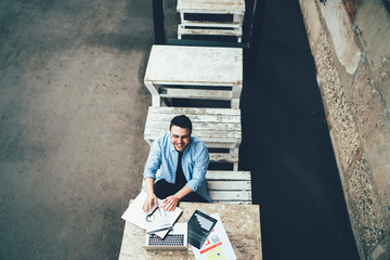 Smiling man working with document and looking at camera at table with laptop in cafe