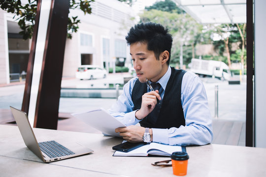 Serious modern Asian businessman looking through documents on street terrace