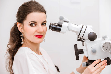 Attractive young female dentist in a white suit uses a microscope at work