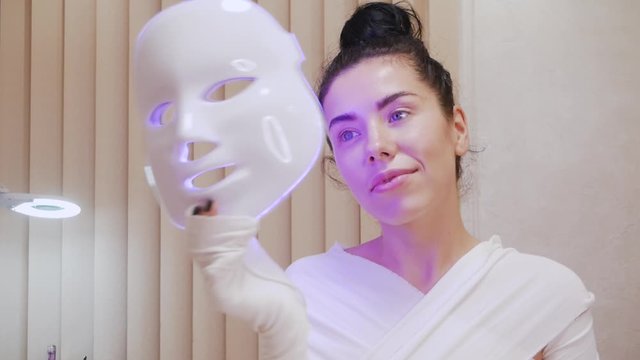Modern technologies of beauty and health. Girl in elastic bandage with LED anti-aging mask for facial skin care in spa slow motion. Woman lying on a sofa in a special mask