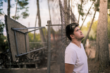 Stock photo of a boy leaning on a fence with a thoughtful expression next to some trees and an abandoned basketball basket