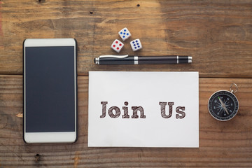 Join Us Words written on white on wooden desk with dice,compass,smart phone and pen.Top view conceptual