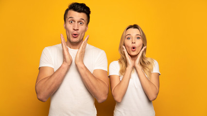Fototapeta premium As like as two peas. A wonderful couple in white t-shirts are posing with their palms near their chins, looking in the camera with surprised facial expressions.