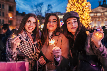 New Year concept. Women friends burning sparklers in Lviv by Christmas tree on street fair. Girls holding shopping bags