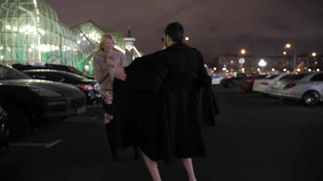 funny prevert in black coat approaches elegant lady and shows bare body on parking lot at night backside view
