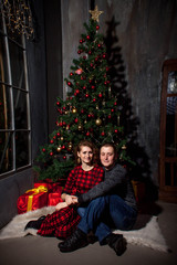 A guy and a girl sit on a white soft rug near a Christmas tree and kiss