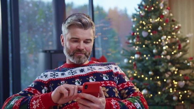 Bearded handsome man using a smartphone near beautiful Christmas tree. Positive caucasian father celebrating holidays at home reading greetings in social network.