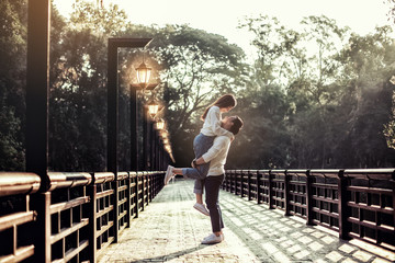 The strong Asian man hold up his girlfriend at the bridge with lamp all the way. There are forest at background.