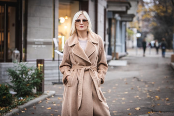 Fototapeta premium Young beautiful blond woman in an autumn coat stands on a city street. Soft focus. Fashionable concept.