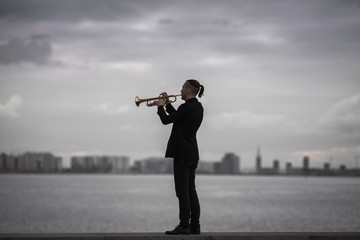 young stylish jazzman playing trumpet on stone seafront against background of the city