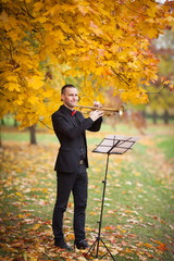young musician in black suit plays  trumpet  in autumn park near music stand with notes among trees 