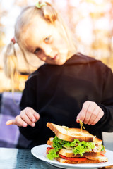 Cute adorable caucasian blond kid girl enjoy eating hamburger with ham and fresh vegeatables at cafe on bright sunny day outdoors. Fast food children unhealthy diet. Snack for having lunch at picnic