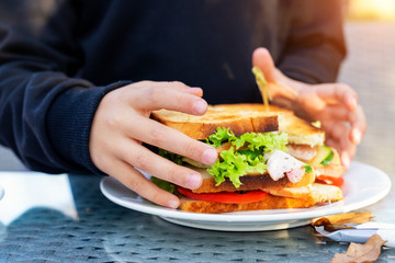 Close-up kid girl hand eating hamburger with ham and fresh vegeatables at cafe on bright sunny day outdoors. Fast food children unhealthy diet. Snack for having lunch at picnic