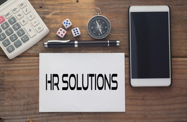 HR SOLUTIONS written on paper,Wooden background desk with calculator,dice,compass,smart phone and pen.Top view conceptual.