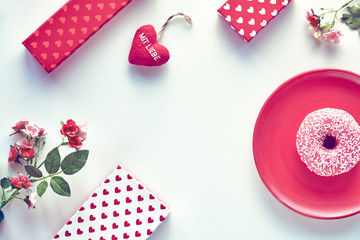 Valentine day flat lay, top view on white table. Geometric background with pussy willow. Gift boxes, pink doughnut on red plate and heart in hand. German text "Mit liebe" on heart means "with love".