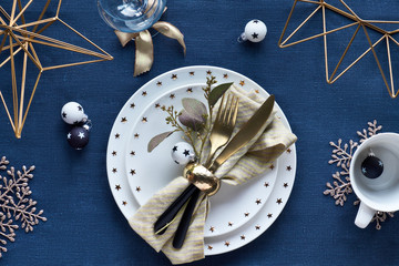 Christmas table setup with white plate and golden utensils and gilded decorations. White and black glass baubles with stars. Flat lay, top view on dark blue linen textile background.