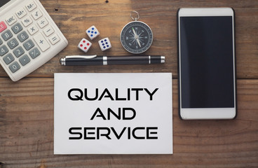 Quality and service written on paper,Wooden background desk with calculator,dice,compass,smart phone and pen.Top view conceptual.