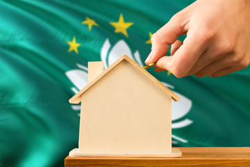 Macao home savings concept. Close-up of hand inserting coin in the wooden house on wooden table with national flag background. Real estate concept.