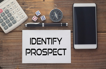 Identify Prospect written on paper,Wooden background desk with calculator,dice,compass,smart phone and pen.Top view conceptual.