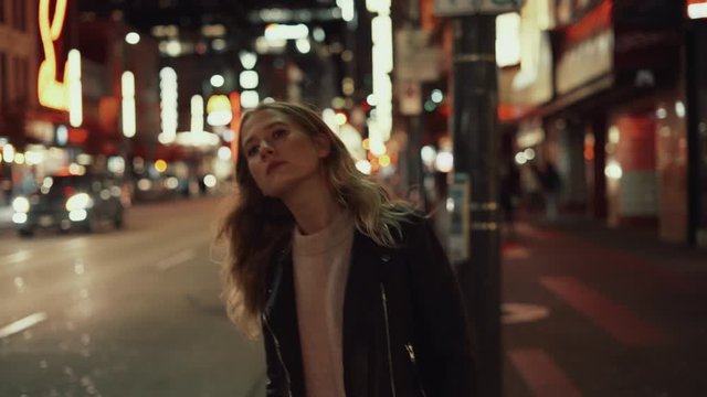 Young blonde woman in jeans and leather jacket posing and walking on streets in downtown after sunset in the evening