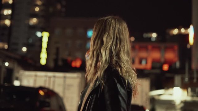 Young blonde woman in jeans and leather jacket posing and walking on streets in downtown after sunset in the evening