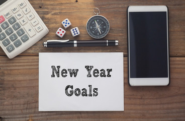 New Year Goals written on paper with wooden background desk with calculator,dice,compass,smart phone and pen.Top view conceptual
