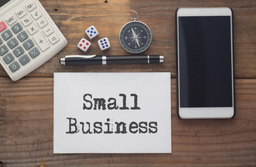 Small Business written on paper,Wooden background desk with calculator,dice,compass,smart phone and pen.Top view conceptual.