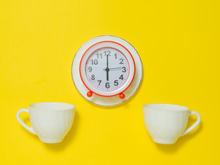 A red alarm clock on a saucer and two coffee cups on a yellow background. Flat lay.