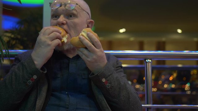 elderly pensioner in fashionable jacket and glasses with gray beard bites two hamburgers in turn. man is very hungry and angry