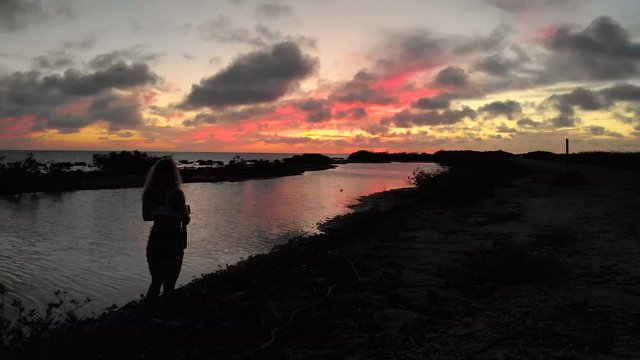 Dolly video of a woman taking pictures of the colorful sky during sunset in front of the calm ocean.
