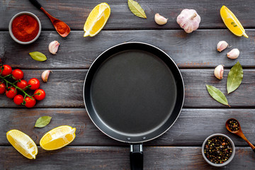 Cooking mockup. Frying pan among spices and vegetables on dark wooden desk top-down copy space