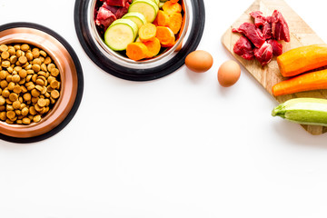 Pet feed ingredients. Raw meat and fresh vegetables near bowl with dry feed on white background top-down copy space