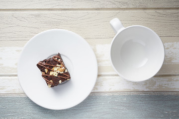 A moist dark chocolate brownie with mix nut on top served on white round plate with empty white cup.