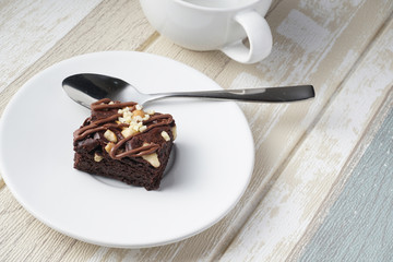 A moist dark chocolate brownie with mix nut on top served on white round plate with silver spoon. A white cup placed next to it.