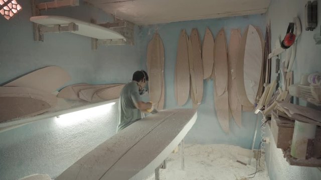 Sequence of shots of Asian craftsman wearing safety respirator sawing surfboard blank lying on workbench using handsaw