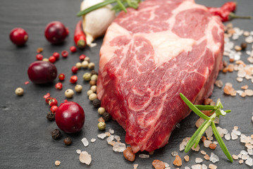 Raw rib eye steak with spices and vegetables. Ingredients for restaurant meal. Fresh meat, salt, rosemary, thyme, chilli, cherry tomatoes, garlic on black stone. Food background.
