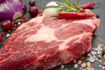 Raw rib eye steak with spices and vegetables. Ingredients for restaurant meal. Fresh meat, salt, rosemary, thyme, chilli, cherry tomatoes, garlic on black stone. Food background.