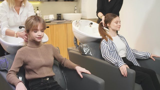 Young caucasian girls enjoy and relax with closed eyes while professional stylists hairdressers wash their heads after successful dyeing in beauty saloon