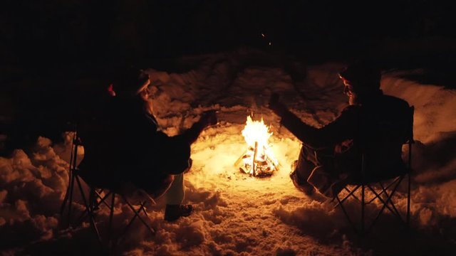 Young couple sitting by the campfire having conversation and drinking in winter at night