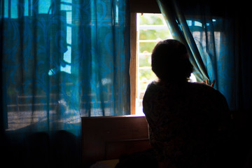 Blurred silhouette of an Indian Bengali brunette woman looking outside by removing the blue curtain of the window in the afternoon. Indian lifestyle.