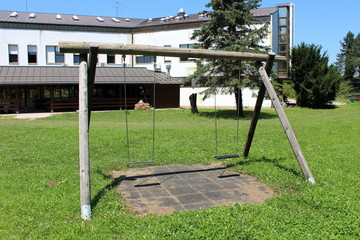 Fototapeta premium Dilapidated old wooden retro vintage outdoor public playground equipment in shape of swing with two seats in abandoned office building backyard surrounded with grass and trees