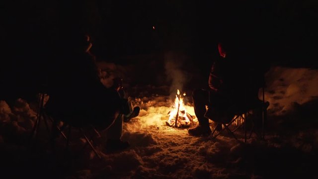 Young couple sitting by the campfire having conversation and drinking in winter at night