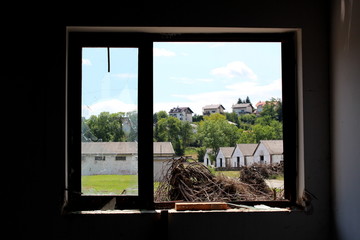 Large and small windows with broken and missing glass at abandoned house overlooking piles of cut down trees and military complex hangars surrounded with dense trees and family houses on warm sunny