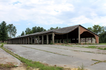 Fototapeta na wymiar Large long hangar building with missing destroyed red brick support walls and unusual bent roof surrounded with paved area mixed with grass and trees at abandoned military complex on cloudy blue sky b